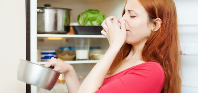 young woman in red holding her nose because of bad smell from food near refrigerator at home